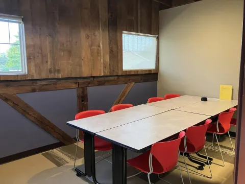 Photograph of Study Room B. Four rectangular tables are pushed together. Seven orange chairs are visible and two windows.