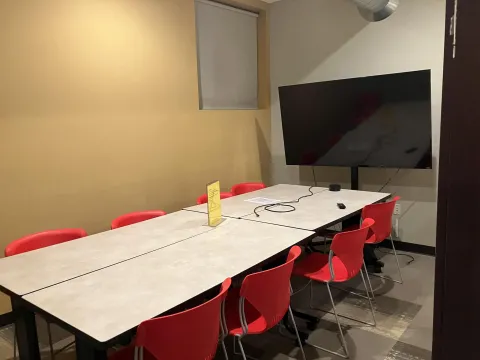 Photograph of Study Room E. Four rectangular tables have been pushed together to form one large table. A smart TV is along the back wall. Eight orange chairs and one window are visible.